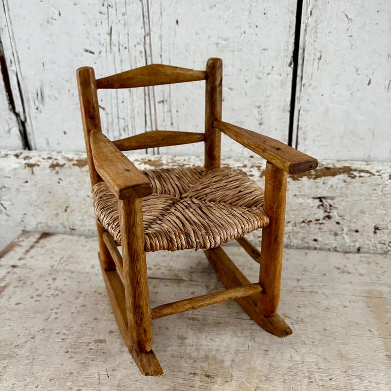 May include: A small, antique wooden rocking chair with a woven straw seat and armrests. The chair has a natural wood finish and is designed for a doll or small child. The chair is approximately 30 cm tall.