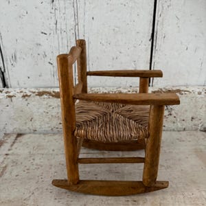 May include: A miniature wooden rocking chair with a woven seat. The chair has a natural wood finish and armrests. The design is simple and rustic, suggesting a vintage or handcrafted origin. It is likely a decorative item or a child's toy, with a charming, old-fashioned aesthetic.