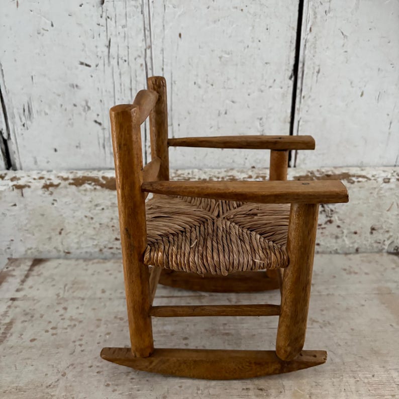 May include: A miniature wooden rocking chair with a woven seat. The chair has a natural wood finish and armrests. The design is simple and rustic, suggesting a vintage or handcrafted origin. It is likely a decorative item or a child's toy, with a charming, old-fashioned aesthetic.