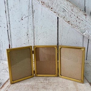 May include: A gold-colored, three-panel picture frame. Each panel has a rectangular shape with a decorative border. The frame is hinged, allowing it to fold. The background is a weathered, white wooden surface.