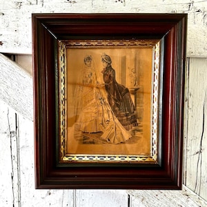 Puede incluir: Impresión en sepia enmarcada que representa a dos mujeres con vestidos de la época victoriana. La impresión está enmarcada en un marco de madera rojo oscuro con un borde interior dorado. La imagen muestra una ilustración detallada de las mujeres en una habitación.