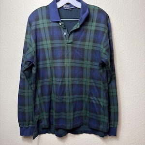 May include: A long-sleeve polo shirt featuring a blue and green plaid pattern. The shirt has a solid blue collar, a three-button placket, and a Polo by Ralph Lauren label at the neckline. The fabric has a textured finish with ribbed cuffs.