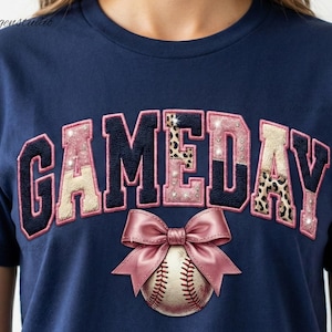 Könnte beinhalten: Marineblaues T-Shirt mit dem Wort "GAMEDAY" im College-Stil, mit rosa und Leopardenmuster-Akzenten. Eine rosa Schleife und ein Baseball befinden sich unter dem Text. Das Shirt ist ein lässiger Stil.