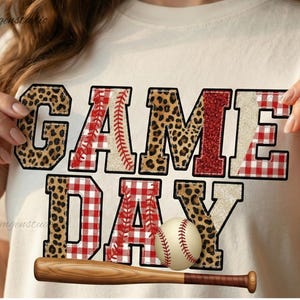 May include: A cream-coloured t-shirt with the words "GAME DAY" in a layered design. The letters feature leopard print, red glitter, and a baseball stitch pattern. A baseball and wooden bat are positioned below the text, creating a sporty theme.
