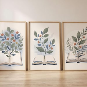 Pode incluir: Um conjunto de três quadros de parede, cada um apresentando um livro aberto com uma ilustração botânica de folhas verdes e pequenas flores laranja crescendo das páginas. As molduras são de madeira clara com um fundo cor de creme.