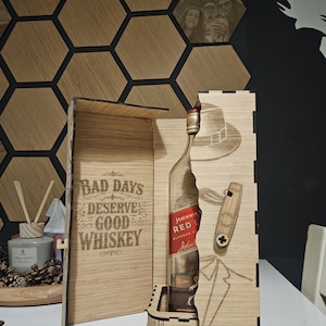 May include: A wooden whiskey gift box with a bottle of Johnnie Walker Red Label. The box is laser-cut with a tuxedo design and the text "BAD DAYS DESERVE GOOD WHISKEY". The bottle is partially visible.
