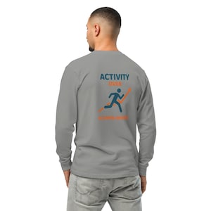 Op de afbeelding: Een grijze longsleeve met de tekst "ACTIVITY OVER ACCOMPLISHING" en een afbeelding van een persoon die een oranje pijl oploopt. De shirt is gemaakt van een zacht materiaal.