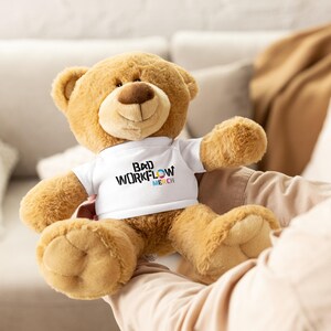 May include: A plush teddy bear with light brown fur, wearing a white t-shirt. The t-shirt has the text "BAD WORKFLOW MERCH" in black, cyan, magenta, and yellow. The bear is sitting with its arms outstretched.