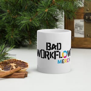 May include: White ceramic mug with the text "BAD WORKFLOW MERCH" in black, blue, yellow, and pink. The mug is cylindrical and sits on a white surface with a dried orange slice and cinnamon sticks.