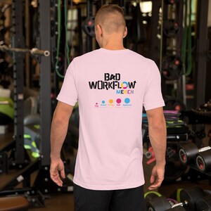 Op de afbeelding: Lichtroze t-shirt met de zwarte tekst "BAD WORKFLOW MERCH" en kleurrijke accenten. Daaronder staat "Say Smart Things Get Applause". Het shirt heeft korte mouwen en is gemaakt van een zacht materiaal.