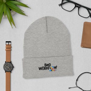 May include: A light grey beanie hat with the text "BAD WORKFLOW MERCH" in black, blue, cyan, and magenta. The hat is folded at the brim. A brown leather watch, black glasses, and a small brown wallet are also visible.