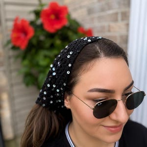 May include: A black crocheted headscarf featuring a triangular pattern with scattered iridescent sequins throughout the fabric. The accessory is worn over the hair and covers the top of the head, showcasing a textured, open-knit construction.