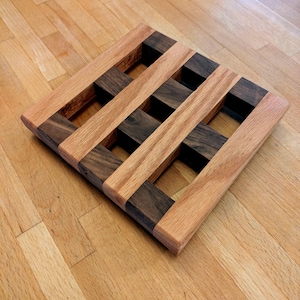 May include: A square wooden trivet with a grid pattern. Alternating light and dark wood blocks create the design. The trivet is made of natural wood and is designed to protect surfaces from hot dishes.
