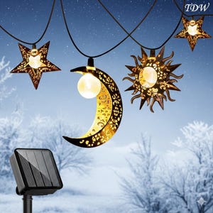 May include: Decorative solar-powered string lights featuring star, sun, and crescent moon shapes. The lights have a warm glow and are suspended from a black wire. A solar panel is visible in the foreground, set against a wintery background.