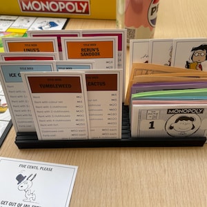Könnte beinhalten: Ein Monopoly-Brettspiel-Set mit Titelkarten, Geld und Spielfiguren. Die Titelkarten tragen Namen wie "Linus's" und "Rerun's Sandbox". Das Spielgeld zeigt eine Cartoonfigur. Das Spiel befindet sich auf einer Holzoberfläche.