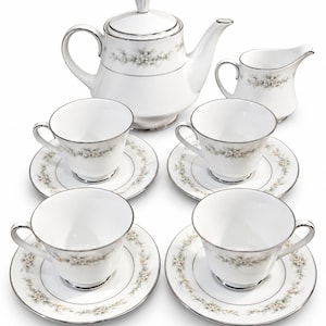May include: A white porcelain tea set comprising a teapot, a creamer, and four teacups with matching saucers. Every item is adorned with a subtle floral design in muted grey and beige shades, finished with a polished silver rim around the edges.