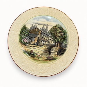 May include: A round ceramic plate featuring a central illustration of a stone house with a wooden balcony, surrounded by trees and flowers. The wide, cream-coloured border is decorated with an embossed scrollwork pattern and a thin brown rim.