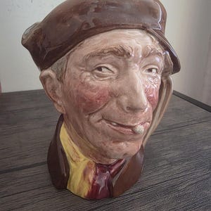 Könnte beinhalten: Keramikfigur eines Männerkopfes mit braunem Barett und Jacke über einem gelben Hemd und roter Krawatte. Der Mann hat rosige Wangen und ein leichtes Lächeln. Die Figur ist wahrscheinlich ein Dekorations- oder Sammlerobjekt.