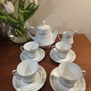 Może przedstawiać: Biały porcelanowy zestaw do herbaty z kwiatowymi detalami, w tym czajniczek, dzbanuszek do śmietanki, cukiernica i cztery filiżanki do herbaty ze spodkami. Zestaw ustawiony jest na drewnianej powierzchni, z wazonem białych tulipanów w tle.