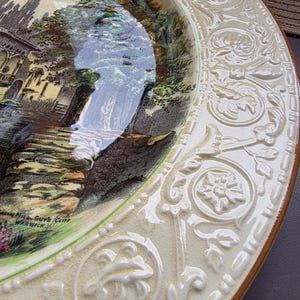 Peut inclure: Assiette décorative vintage crème avec une scène de paysage d'une cascade et de bâtiments. L'assiette a une bordure verte et un motif floral en relief sur le bord. Le texte "Guy's Cliff Warwick" est visible.
