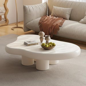 May include: A modern, white, kidney-shaped coffee table with three cylindrical legs. The table is accessorized with a small bowl of green apples, two decorative vases, and a stack of books. The table sits on a neutral-colored rug.