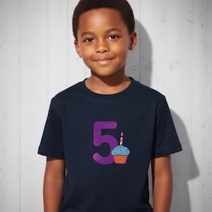 Puede incluir: Camiseta azul marino con el n&uacute;mero 5 morado y un gr&aacute;fico de cupcake. La magdalena tiene glaseado azul y una envoltura naranja, con una vela encendida. La camiseta la lleva un ni&ntilde;o.