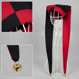 Op de afbeelding: Een narrenkap met een zwart-rood ruitpatroon op de kroon en twee lange, puntige stoffen uiteinden die naar beneden hangen. Elk uiteinde is afgewerkt met een klein, goudkleurig metalen belletje dat aan de punt van de stof is bevestigd.