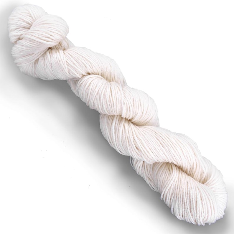Puede incluir: Una madeja de hilo color blanco hueso retorcida en una bobina larga y continua. El hilo presenta una textura suave de m&uacute;ltiples cabos con un color crema claro consistente en toda la longitud de la fibra, sobre un fondo blanco liso.