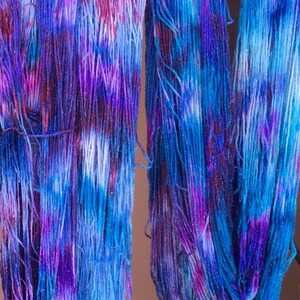 Puede incluir: Dos madejas de hilo te&ntilde;ido a mano con una paleta de colores variados en tonos azul vibrante, morado intenso y magenta. Las fibras del hilo est&aacute;n entremezcladas con sutiles hilos de purpurina met&aacute;lica que captan la luz en las hebras.