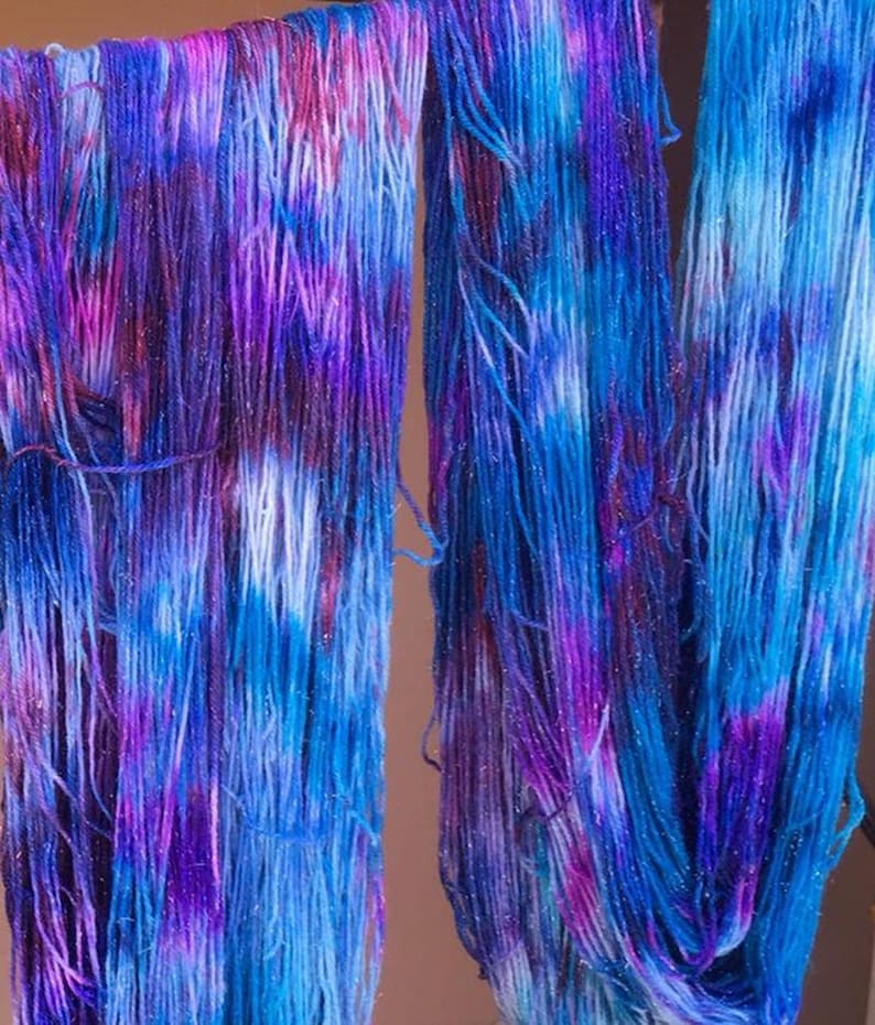 Puede incluir: Dos madejas de hilo te&ntilde;ido a mano con una paleta de colores variados en tonos azul vibrante, morado intenso y magenta. Las fibras del hilo est&aacute;n entremezcladas con sutiles hilos de purpurina met&aacute;lica que captan la luz en las hebras.