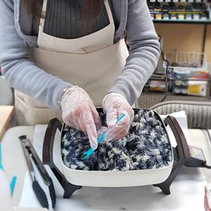Puede incluir: Una sart&eacute;n el&eacute;ctrica blanca que contiene una masa de hilo con un patr&oacute;n moteado en blanco y negro. El hilo es manipulado con una peque&ntilde;a herramienta de pl&aacute;stico azul por manos con guantes desechables transparentes. Unas pinzas de metal est&aacute;n cerca.
