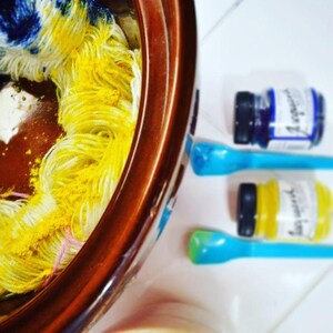Puede incluir: Un cuenco de cer&aacute;mica marr&oacute;n que contiene hilo blanco saturado con polvo de tinte amarillo y azul. Junto al cuenco hay dos frascos de pl&aacute;stico peque&ntilde;os con tapas negras que contienen tinte azul y amarillo, cada uno con una cuchara de pl&aacute;stico azul claro.