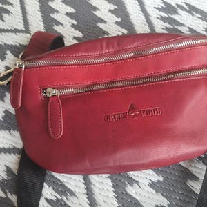 Puede incluir: Un cinturón de cuero rojo con cremalleras plateadas y una correa negra ajustable. La bolsa tiene el texto en relieve "GREEN WOOD" e incluye una pequeña bolsa a juego. La bolsa está sobre una superficie estampada.
