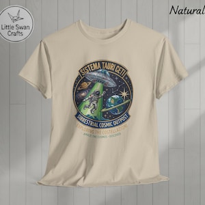 Può includere: T-shirt beige con grafica circolare raffigurante un astronauta, un UFO e pianeti. La grafica include il testo "SISTEMA TAURI CETI" e "SIRRESTRIAL COSMIC OUTPOST". La maglietta ha anche il testo "EXPLORING THE COSTELLAZIONI" e "AMAZE THE COSMOS - DISCOVER".