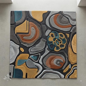 Può includere: Un tappeto quadrato caratterizzato da un design astratto e materico con sezioni intagliate in rilievo. Il motivo è composto da forme organiche e fluide nei toni del grigio antracite, giallo senape, arancione bruciato, grigio chiaro e blu ottanio.