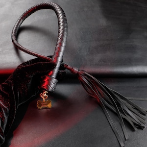 Puede incluir: Látigo de cuero negro con mango trenzado y borla de finas tiras de cuero. El mango lo sujeta una mano enguantada. Una pequeña etiqueta dorada con la palabra "Qutek" está sujeta al mango.