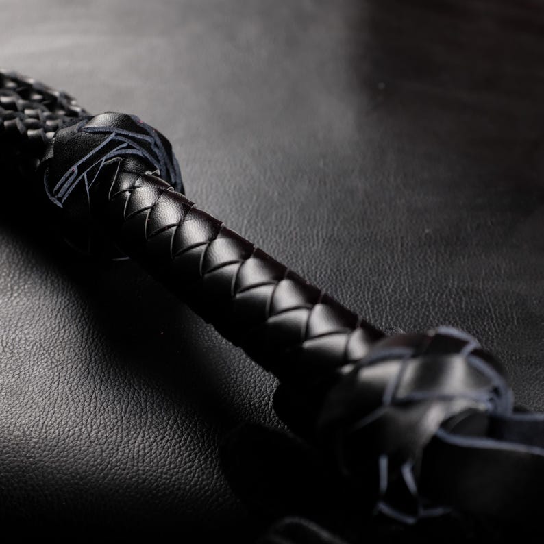 May include: Close-up of a black leather whip. The handle is tightly braided, transitioning to a wider, woven section. The leather appears smooth and well-crafted, with a dark, glossy finish. The background is a textured, dark surface.