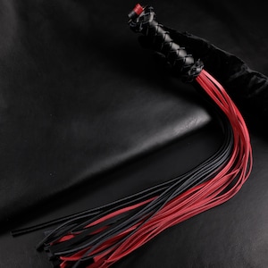 Puede incluir: Látigo de cuero negro y rojo. El mango es de cuero negro trenzado con un acento rojo. El látigo tiene un grupo de tiras de cuero rojas y negras. El fondo es negro.