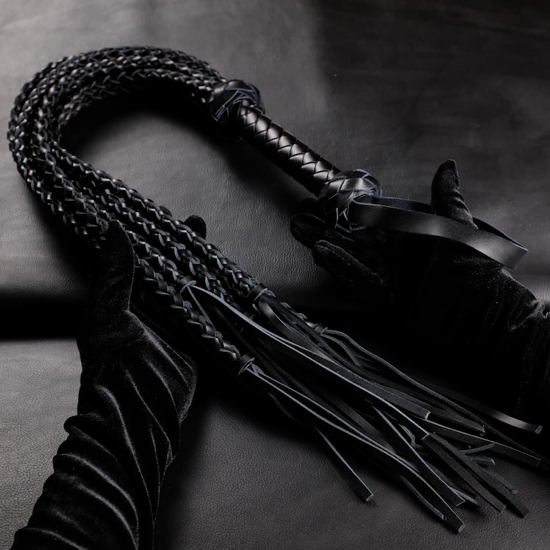 May include: A black leather whip with a braided handle and multiple braided strands. The whip is held by a person wearing black gloves. The background is a dark, textured surface.