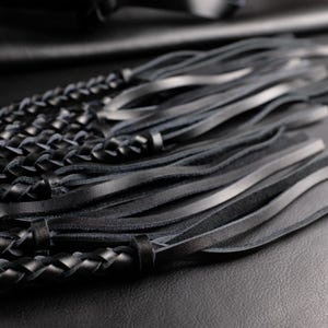 May include: Close-up of a black leather accessory featuring braided and fringed elements. The braided section is tightly woven, while the fringe consists of long, slender strips. The leather appears smooth and has a slight sheen, suggesting a high-quality material.