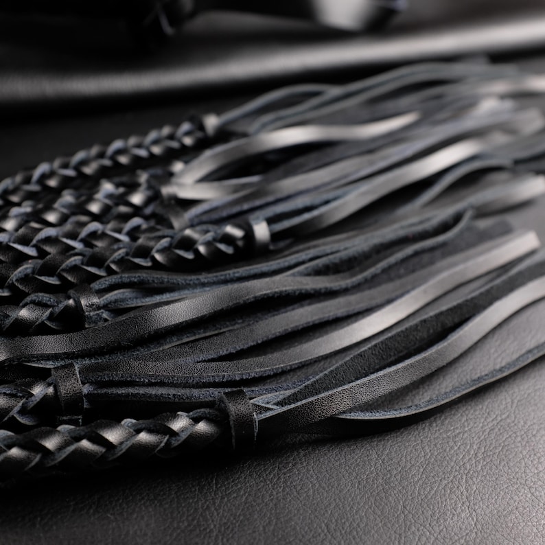 May include: Close-up of a black leather accessory featuring braided and fringed elements. The braided section is tightly woven, while the fringe consists of long, slender strips. The leather appears smooth and has a slight sheen, suggesting a high-quality material.