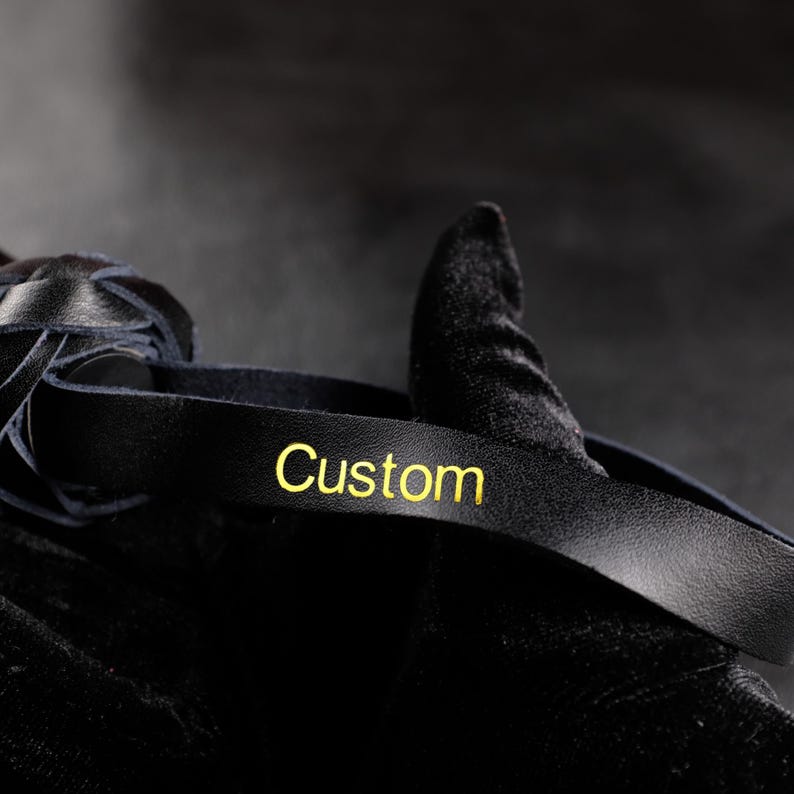 May include: Close-up of a black leather strap with the word "Custom" in gold lettering. The strap is curved and appears to be part of a larger item, possibly a handle or accessory. The background is dark and out of focus.