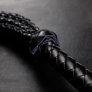 May include: Close-up of a black leather whip with a braided handle and a knotted end. The whip's texture is visible, showcasing the craftsmanship. The image is set against a dark background, highlighting the details of the leatherwork.