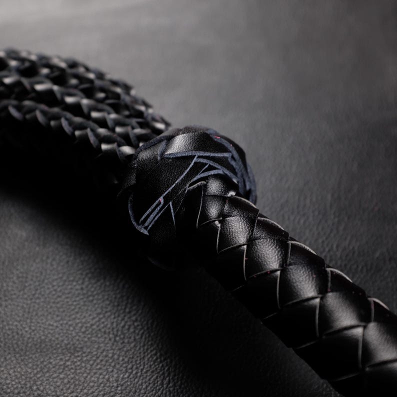May include: Close-up of a black leather whip with a braided handle and a knotted end. The whip's texture is visible, showcasing the craftsmanship. The image is set against a dark background, highlighting the details of the leatherwork.