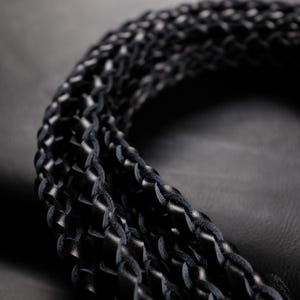 May include: Close-up of a black leather braided cord, likely for a belt or strap. The cord is tightly woven, with a textured surface. The background is a dark, solid color, emphasizing the detail of the braided leather.