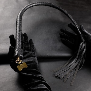 Puede incluir: Látigo de cuero negro con mango trenzado y borla de tiras de cuero. Un dije dorado en forma de hueso de perro cuelga del mango. El látigo es sostenido por manos enguantadas sobre un fondo negro.
