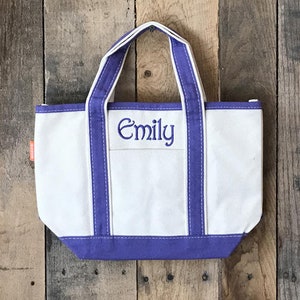 以下が含まれることがあります： 紫色の縁取りと正面に「Emily」と刺繍された白いキャンバス地のトートバッグです。