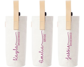 May include: Three white canvas wine bottle bags with tan leather handles. The bags are personalized with the names "Kayla", "Amelia", and "Jessin" in purple script. The words "Bride" and "Bridesmaid" are also printed on the bags.