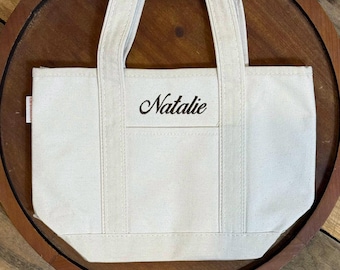May include: A small off-white canvas tote bag featuring two sturdy handles and a front pocket. The name Natalie is embroidered in a black script font on the front center of the bag. A small white fabric tag is visible on the left side seam.