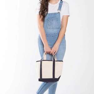 Puede incluir: Una mujer lleva un vestido de peto vaquero azul claro con una camiseta blanca y sandalias de esparto a rayas blancas y negras. Lleva un bolso de mano de lona blanca con ribetes negros.